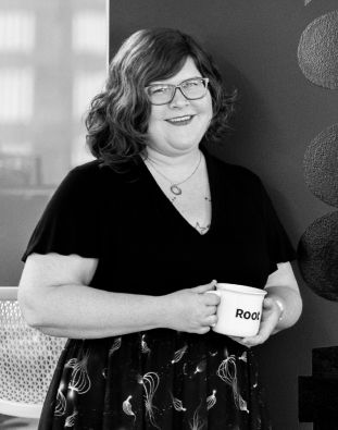 A designer with wavy hair and glasses holds a coffee mug that says “Root.”